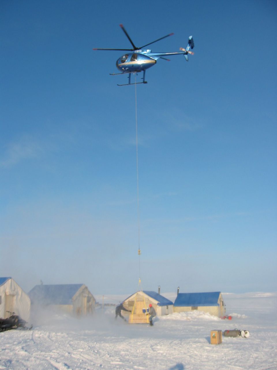 Blue helicopter hovers over a snowy landscape, lifting a small yellow structure with a cable. Several tents and buildings are visible