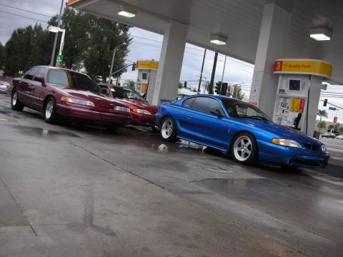 1998 Ford Crown Victoria - Two cars, a blue Ford Mustang and a red Ford Crown Victoria, are at a gas station under a cloudy sky.