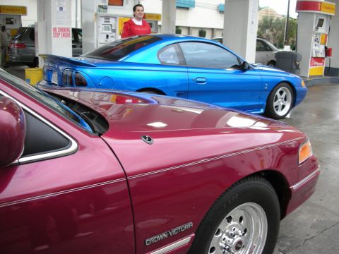 1998 Ford Crown Victoria - A red 1998 Ford Crown Victoria is parked next to a blue Mustang at a gas station.