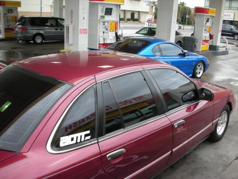 1998 Ford Crown Victoria - Two cars, a maroon sedan and a blue coupe, are at a gas station.