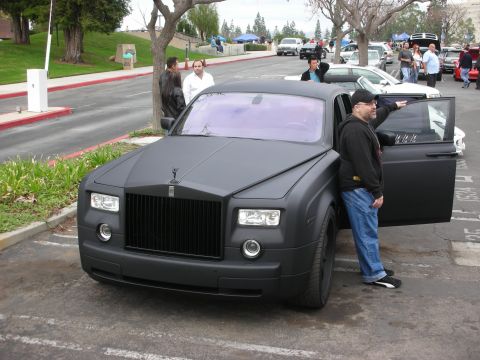 1998 Ford Crown Victoria - A matte black Rolls-Royce Phantom is parked in a lot with other vehicles and people around.