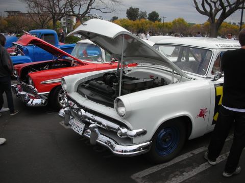 1998 Ford Crown Victoria - A white classic car with its hood open is parked next to a red car and a blue truck.