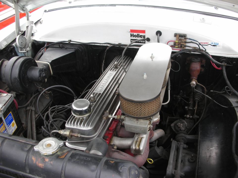 Engine bay showing a V8 engine with Holley carburetor and air cleaner. Radiator and battery visible