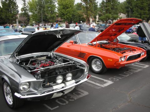1998 Ford Crown Victoria - Two classic cars, one orange and one silver, are parked side-by-side with their hoods open at an outdoor car show.