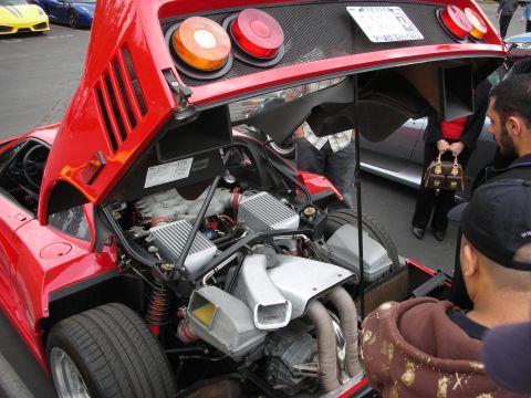 1998 Ford Crown Victoria - The engine bay of a red sports car is open, revealing its twin turbochargers and exhaust system, with onlookers gathered around.