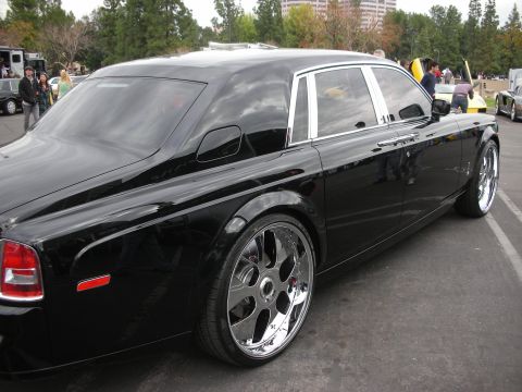 1998 Ford Crown Victoria - A black luxury sedan with custom chrome wheels is parked in a lot, with other vehicles and people in the background.