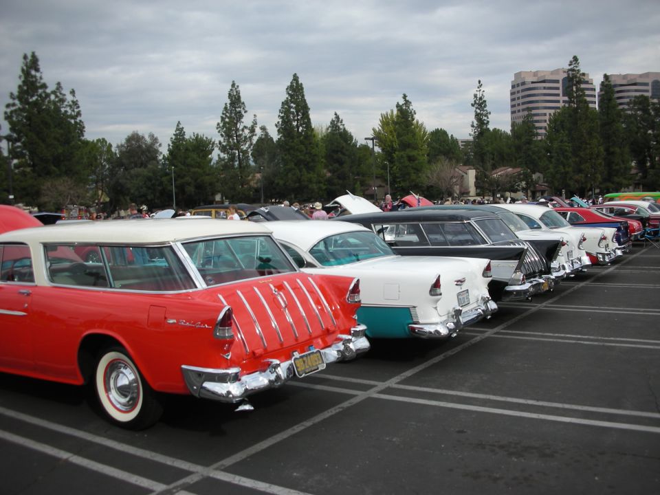 Line of classic cars parked in lot, including a red 1955 station wagon and a black hearse