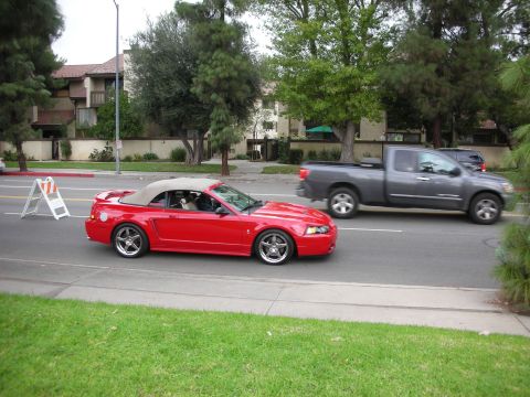 1998 Ford Crown Victoria - A red convertible Mustang is seen driving on a street next to a gray pickup truck.