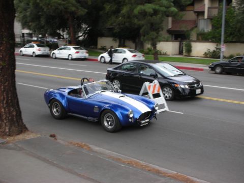 1998 Ford Crown Victoria - A blue convertible sports car with white racing stripes drives down a street, passing other vehicles and oncoming vehicles.