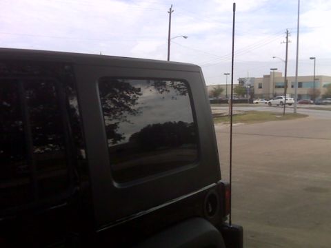 2008 Jeep Wrangler - The rear passenger side of a black Jeep Wrangler is shown parked on a dirt lot with a building and cars in the background. The reflection i…