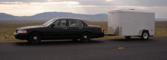Black Ford Crown Victoria towing a white cargo trailer on a road with mountains.