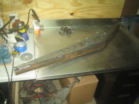 1990 Ford Bronco - Photograph of a fabricated steel suspension/frame member with lightening holes resting on a shop table, not a document.