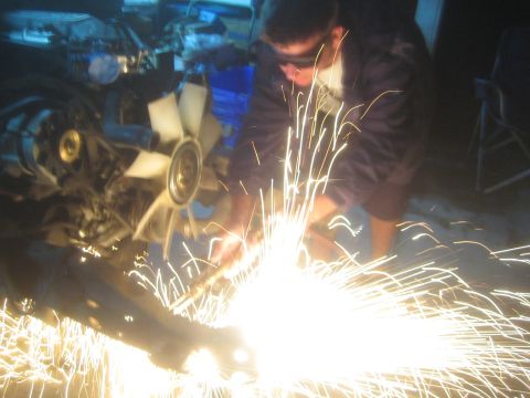 1990 Ford Bronco - Blurry photograph of grinding work on a vehicle frame with heavy sparks, not a document.