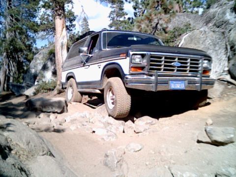 1985 Ford Bronco - This Ford Bronco is navigating a rough, rocky terrain with trees and boulders in the background.