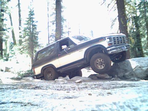 1985 Ford Bronco - A two-tone Ford Bronco navigates rocky terrain in a wooded area.