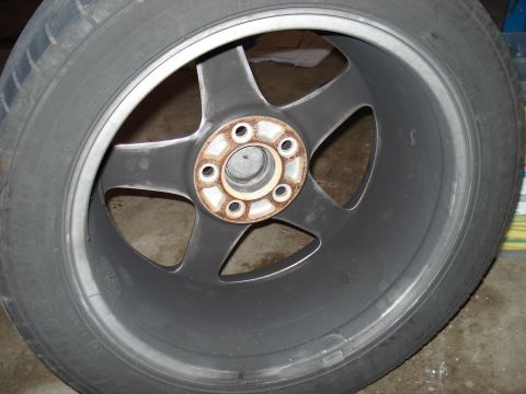 1998 Ford Taurus - A close-up view of a dark gray alloy wheel with five spokes, showing rust around the lug nut holes. The tire is mounted on the wheel.