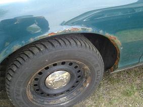 2003 Ford Crown Victoria - A close-up side view of the front passenger-side fender and wheel well of a dark teal vehicle, showing significant rust around the wheel op…