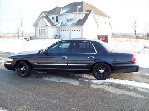 1999 Mercury Grand Marquis - Driver-side profile of a dark navy blue Mercury Grand Marquis four-door sedan on black steel wheels with no hubcaps, parked at the curb of…