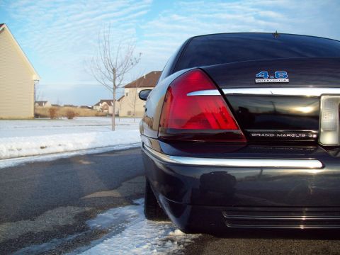 1999 Mercury Grand Marquis - Low close-up of the driver-side rear corner of a dark blue Mercury Grand Marquis GS, showing the wraparound red tail lamp, chrome bumper st…