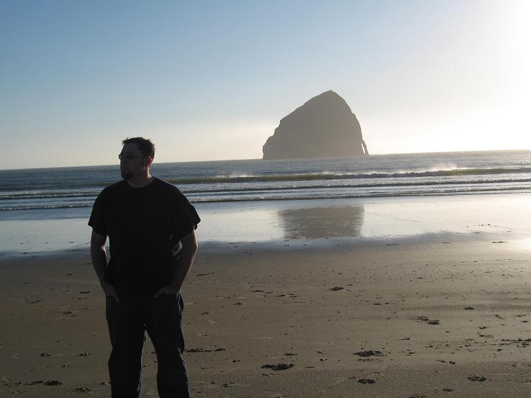 Guy standing on a beach, looking out at the ocean with a large rock formation offshore