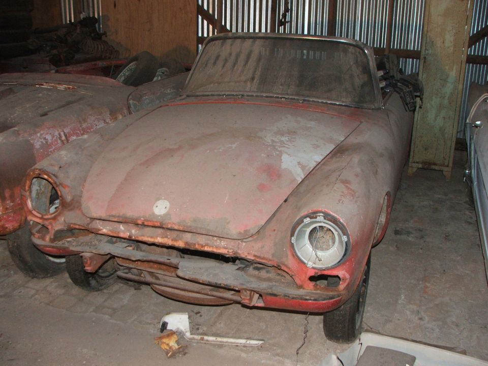 Dusty red convertible with missing headlights and grille, parked next to another car in a garage