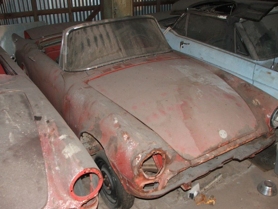 Dusty red convertible sports car with missing headlights and grille, parked indoors next to a blue car