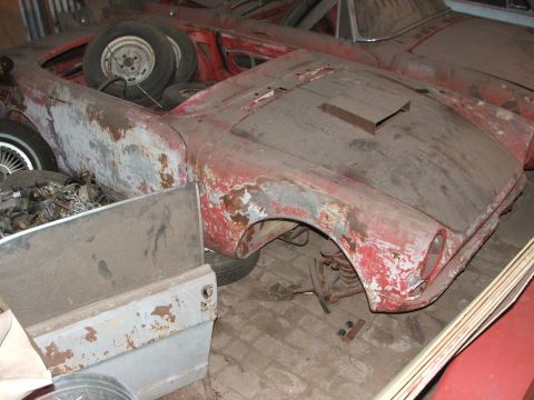 1965 Ford Mustang - A weathered red convertible, possibly a 1965 Sunbeam Alpine, sits in storage with spare tires piled in the passenger area.