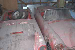 1965 Ford Mustang - Two weathered red convertible cars sit in storage, one with spare wheels piled on its hood.