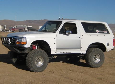 1996 Ford Bronco - A white, lifted 1996 Ford Bronco is parked on a dirt lot with RVs and mountains in the background. The vehicle has large off-road tires, a…