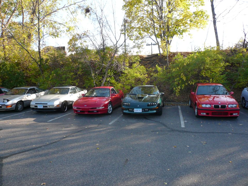 A line of sports cars parked in a lot, including a red Ford Probe, green Camaro, and red BMW.