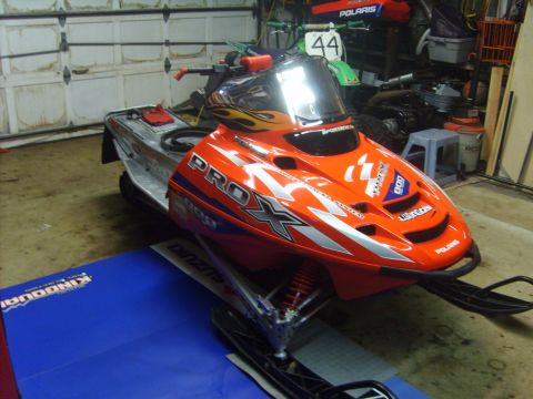 1995 Ford Bronco - A bright orange and silver Polaris PRO-X snowmobile is parked indoors on a blue mat. The snowmobile has flame graphics on the windshield an…