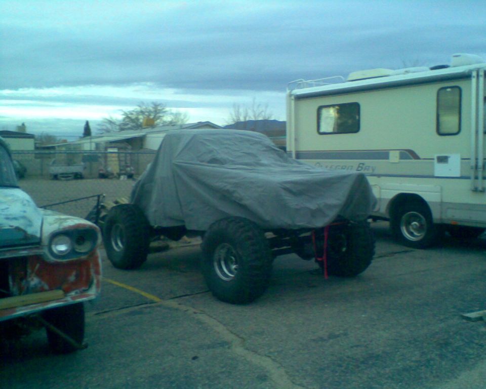 Off-road vehicle covered by tarp next to RV, with part of another vehicle on the left.