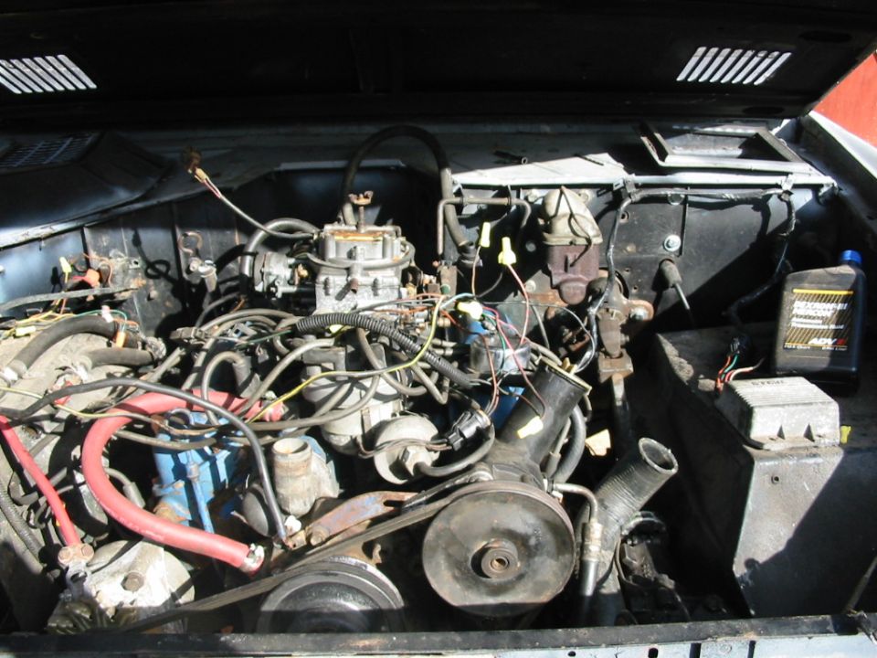 Engine bay of a 1977 Ford Bronco showing carbureted V8 with blue block, hoses, wiring and power steering pump