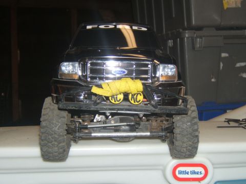 1988 Ford Bronco - Front view of a black Ford F-series truck model with a chrome grille, yellow tow strap, and KC lights. The vehicle is sitting on a white su…