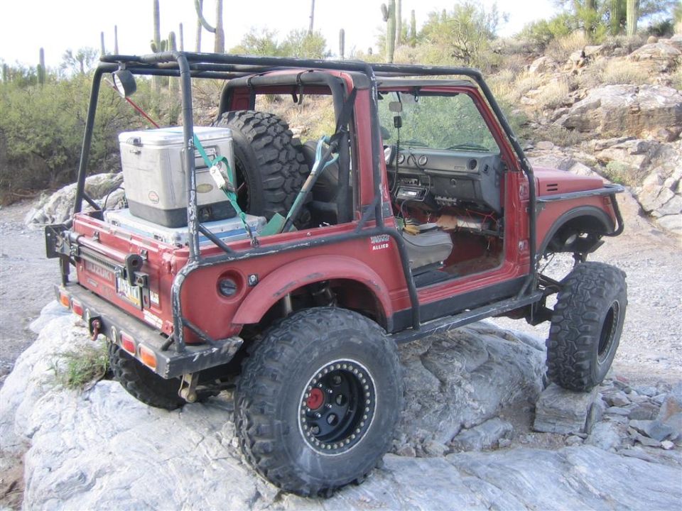 Red 1987 Suzuki Samurai off-roading over rocks with a roll cage and cooler.