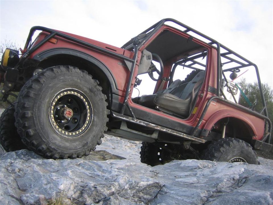 Red modified Suzuki Samurai with large tires and roll cage on rocks.