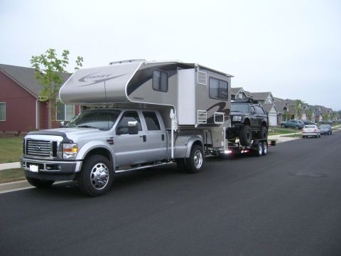 1996 Ford Bronco - A silver Ford F-series Super Duty truck with a camper shell is towing a trailer with a lifted black SUV. The truck and trailer are parked o…
