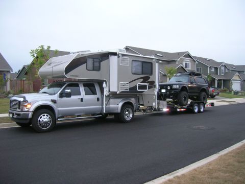 1996 Ford Bronco - A silver Ford F-series truck with a "Host" brand camper shell is towing a trailer carrying a lifted black Ford Bronco. The vehicles are par…