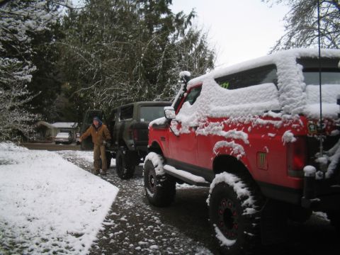 1989 Ford Bronco - A person stands next to a dark green SUV covered in snow, with a red SUV also covered in snow parked in front of it. The vehicles are parke…