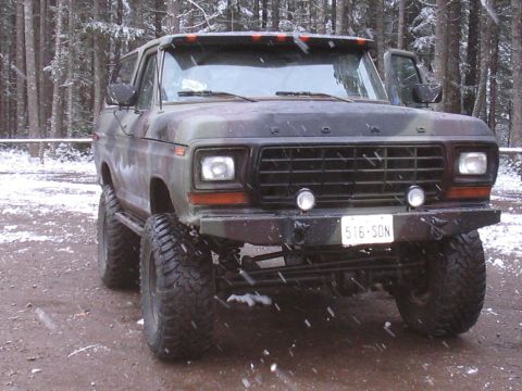 1989 Ford Bronco - A lifted, camouflage-painted 1980s Ford Bronco is parked on a dirt road in a snowy forest. The front of the vehicle, including its large ti…