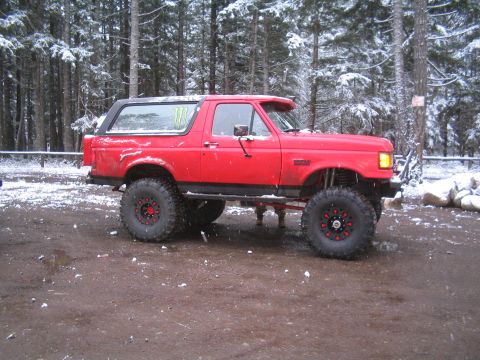 1989 Ford Bronco - A lifted red Ford Bronco is parked on a muddy, snow-dusted ground in a forest. The vehicle has large off-road tires with red accents on the…