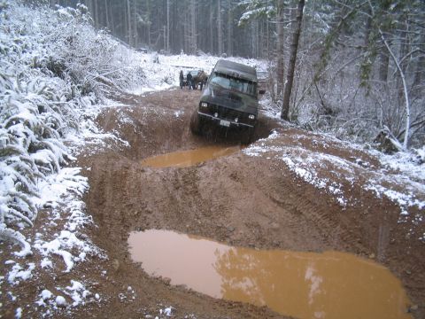 1989 Ford Bronco - A dark-colored SUV navigates a muddy trail with snow on the surrounding trees and ground. The vehicle is partially submerged in a large mud…