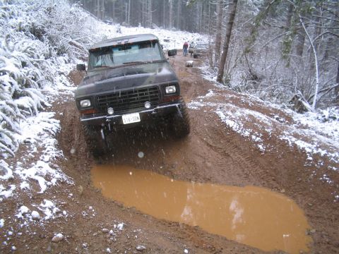 1989 Ford Bronco - A dark-colored Ford Bronco navigates a muddy, snow-covered trail in a forest setting. The vehicle is angled down a steep incline, with its…