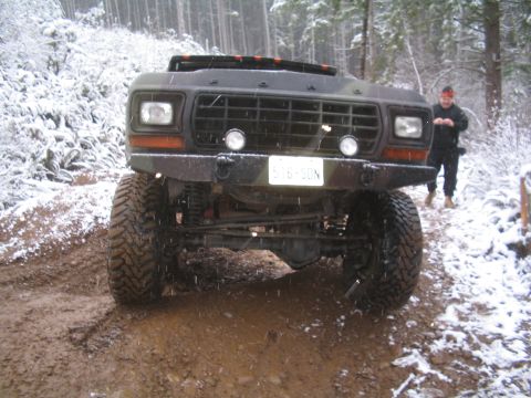 1989 Ford Bronco - A lifted, dark colored Ford Bronco is parked on a muddy, snow-dusted trail in a wooded area. A person in a red headband and dark clothing s…