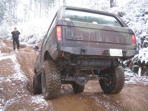 1989 Ford Bronco - The rear driver's side of a dark-colored Ford Bronco is shown angled uphill on a muddy, snow-dusted trail. The vehicle has aggressive off-r…