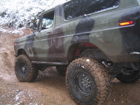 1989 Ford Bronco - A camouflage-painted Ford Bronco is shown from the rear quarter angle on a muddy, snowy trail. The vehicle has large, knobby tires and appe…