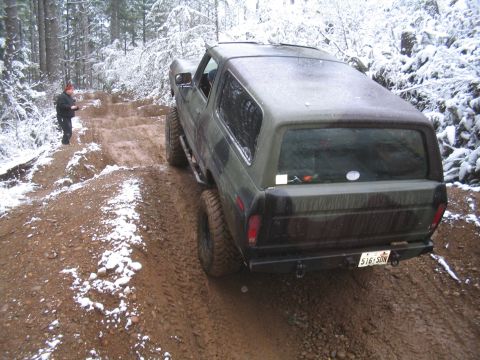1989 Ford Bronco - A dark green camouflage Ford Bronco is driving on a muddy, snow-dusted trail in a forest.