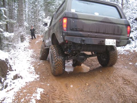 1989 Ford Bronco - The rear view of a camouflage Ford Bronco is shown driving through a muddy, snow-covered forest road. The vehicle has large off-road tires…