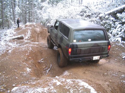 1989 Ford Bronco - A camouflage Ford Bronco is navigating a muddy, snow-dusted off-road trail in a forest. The vehicle is seen from a rear three-quarter angle…