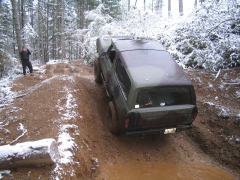 1989 Ford Bronco - A dark green Ford Bronco is driving through a muddy, snow-covered forest trail. The vehicle is viewed from the rear three-quarter angle as…